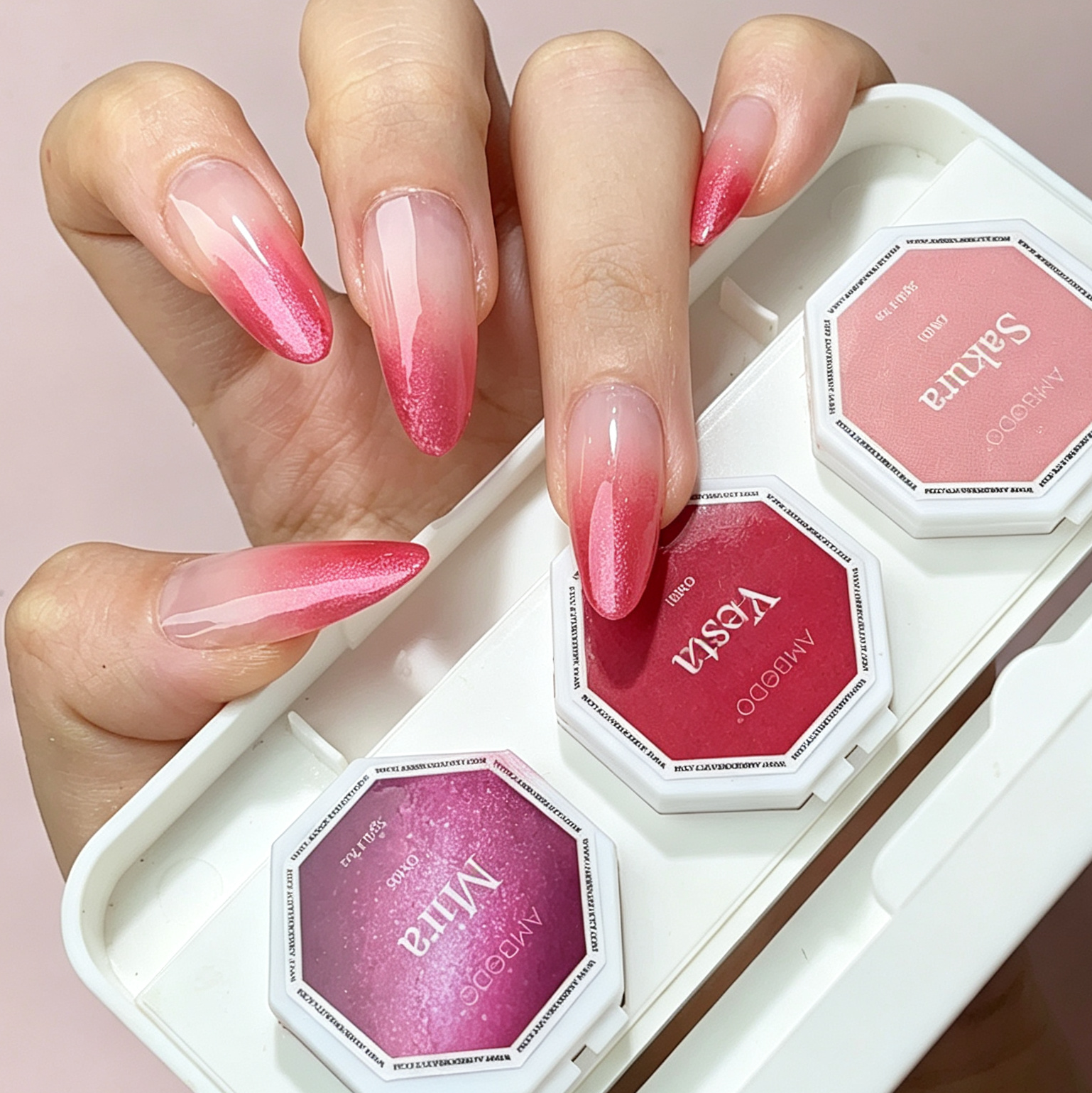 A hand with long pink ombre nails holds a white hexagonal makeup palette containing three pink and magenta cosmetic pans. The background is a soft light pink.